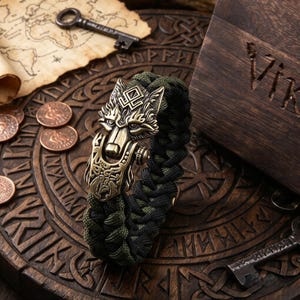 May include: A bracelet featuring a braided olive green and black cord band with a metallic bronze-toned clasp shaped like a stylized wolf head. The clasp displays intricate engraved patterns and a geometric symbol on the wolf's forehead.