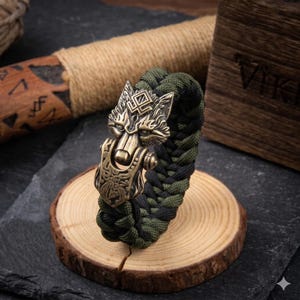 May include: A braided bracelet with a bronze wolf head clasp. The bracelet is made of green and black cord and rests on a small wooden slice. The wolf head clasp features intricate details and a vintage finish.