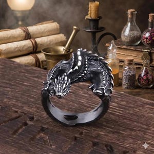 May include: A silver dragon ring with intricate detailing. The dragon is black and white, with its head and body curving around the ring. The ring is displayed on a wooden surface, with other fantasy-themed items in the background.