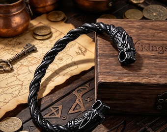 May include: A black metal cuff bracelet featuring a twisted rope design and two stylized animal head terminals. The bracelet is open-ended and displays intricate engraved details on the heads, showcasing a dark, polished finish.