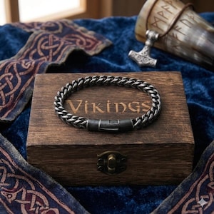 May include: A dark silver-toned metal chain bracelet with a rectangular clasp, resting on a rectangular wooden box featuring the word VIKINGS engraved on the lid. The box has a metal latch and is positioned near a metal hammer pendant.