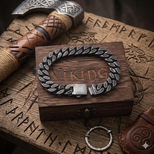 Puede incluir: Un brazalete vikingo plateado con un diseño de eslabones de cadena, colocado sobre una caja de madera con la palabra "Viking" grabada. También se ven un hacha y un anillo.