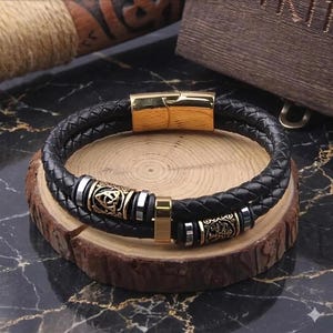 May include: A double-strand black braided leather bracelet featuring a polished gold-tone magnetic clasp, a central gold-tone rectangular spacer, and two cylindrical gold-tone beads with intricate embossed knotwork patterns and silver-tone accents.