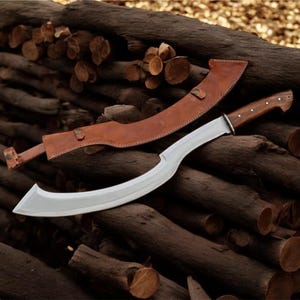 May include: A curved, silver-bladed knife with a wooden handle and a brown leather sheath. The knife is lying on a pile of cut logs. The sheath has a strap and buckle closure.