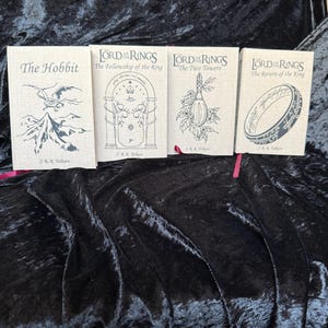 May include: Four hardcover books from the Lord of the Rings and The Hobbit series. The books have a beige linen texture with black illustrations and titles. Titles include "The Hobbit", "The Fellowship of the Ring", "The Two Towers", and "The Return of the King".