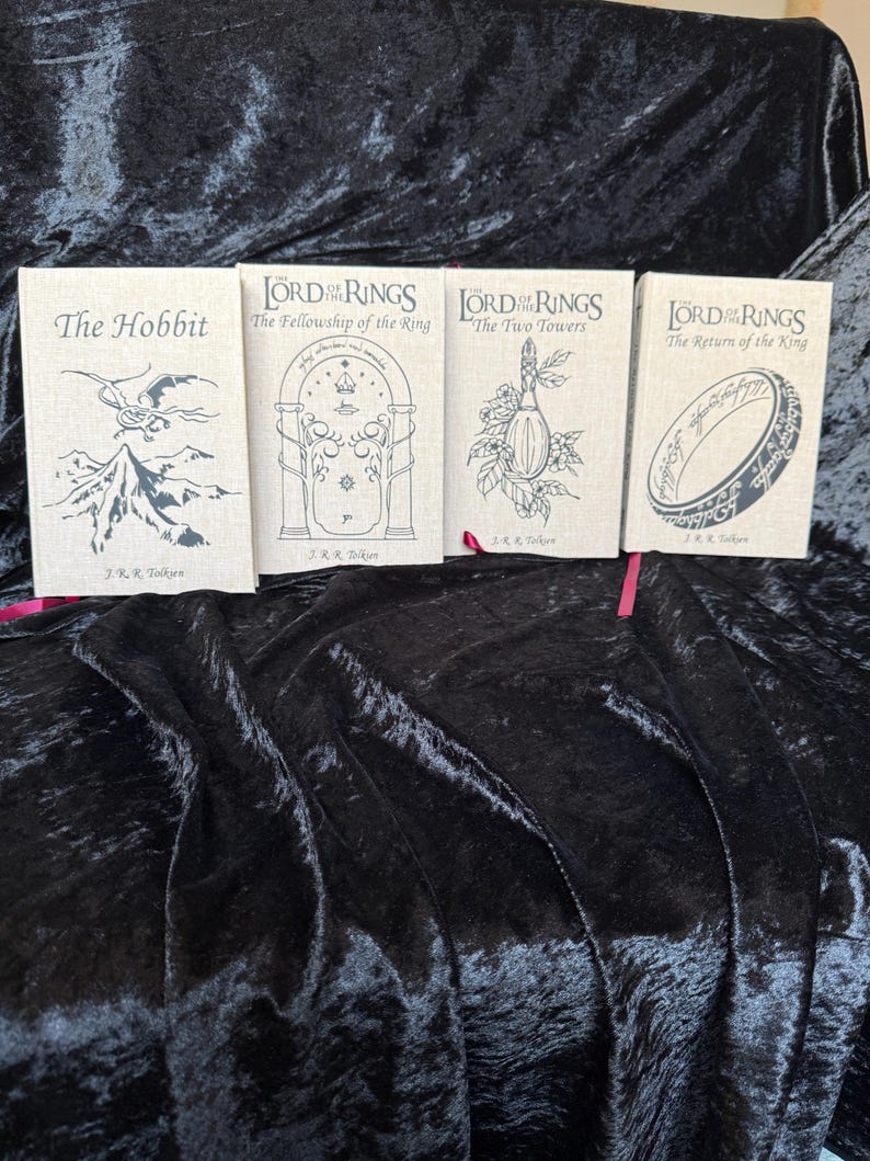 May include: Four hardcover books from the Lord of the Rings and The Hobbit series. The books have a beige linen texture with black illustrations and titles. Titles include "The Hobbit", "The Fellowship of the Ring", "The Two Towers", and "The Return of the King".