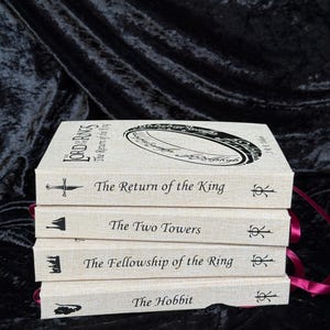 May include: A stack of four books with off-white covers and burgundy ribbon bookmarks. The titles include "The Hobbit", "The Fellowship of the Ring", "The Two Towers", and "The Return of the King". Each book has a unique illustration and title printed in black.