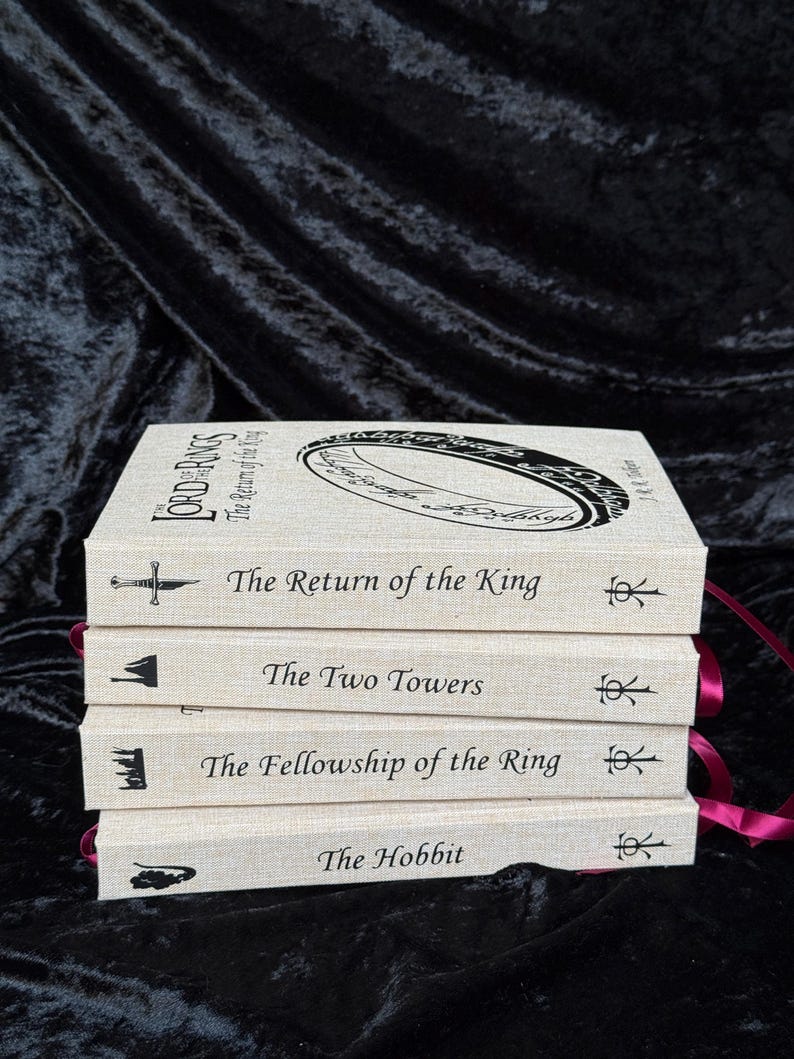 May include: A stack of four books with off-white covers and burgundy ribbon bookmarks. The titles include "The Hobbit", "The Fellowship of the Ring", "The Two Towers", and "The Return of the King". Each book has a unique illustration and title printed in black.