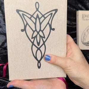 May include: A beige book with a black graphic design on the cover. The design is a stylized emblem. A second book is partially visible in the background. A pink ribbon is attached to the book.