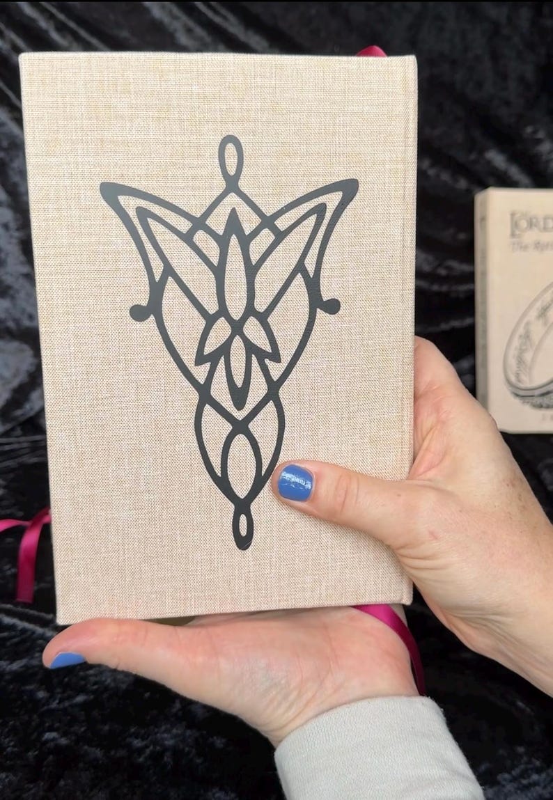 May include: A beige book with a black graphic design on the cover. The design is a stylized emblem. A second book is partially visible in the background. A pink ribbon is attached to the book.