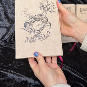 May include: A hardcover book with a beige linen cover featuring a black illustration of a hobbit hole with a tree and smoke. The book has a pink ribbon bookmark. Other books are visible in the background.