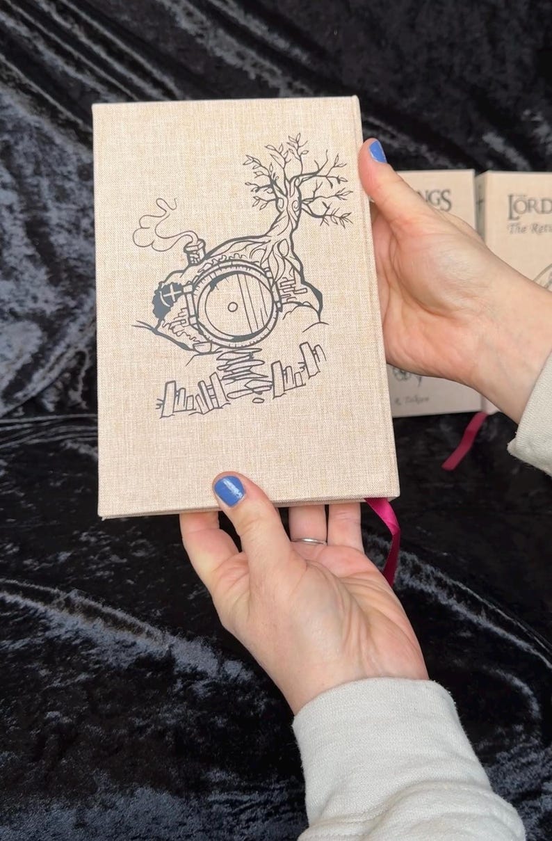 May include: A hardcover book with a beige linen cover featuring a black illustration of a hobbit hole with a tree and smoke. The book has a pink ribbon bookmark. Other books are visible in the background.