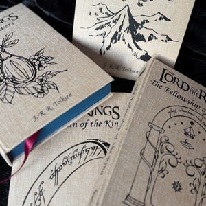 May include: A collection of J.R.R. Tolkien's "The Lord of the Rings" books. The book covers are a light tan color with black illustrations and text. The books have blue and red ribbon bookmarks.
