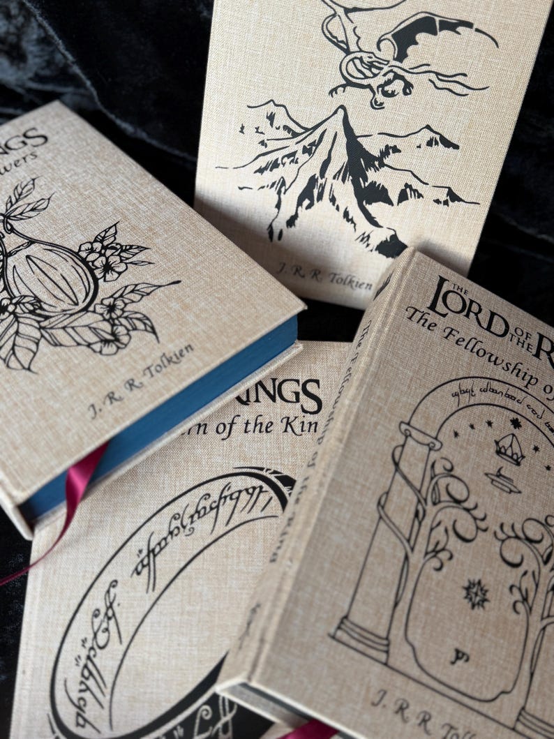 May include: A collection of J.R.R. Tolkien's "The Lord of the Rings" books. The book covers are a light tan color with black illustrations and text. The books have blue and red ribbon bookmarks.