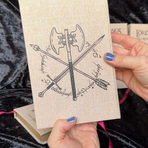 May include: A beige book cover with a black illustration of crossed weapons, including an axe, sword, arrow, and spear. The cover has text in a stylized font. A pink ribbon bookmark is visible. The book is held by a person.