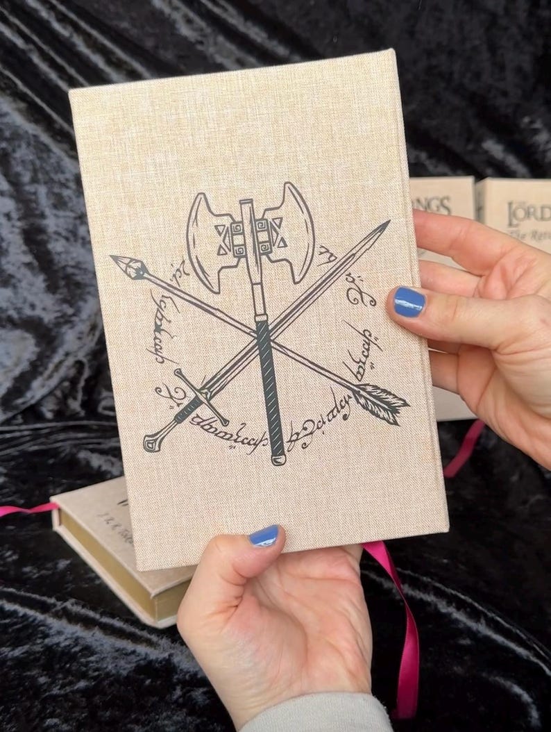 May include: A beige book cover with a black illustration of crossed weapons, including an axe, sword, arrow, and spear. The cover has text in a stylized font. A pink ribbon bookmark is visible. The book is held by a person.
