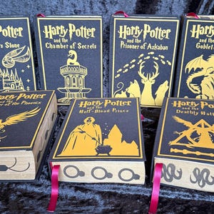 May include: A collection of six decorative book boxes featuring the "Harry Potter" series titles. Each box has a black cover with gold lettering and illustrations, including the Sorcerer's Stone, Chamber of Secrets, and Deathly Hallows. Red ribbon bookmarks are visible.