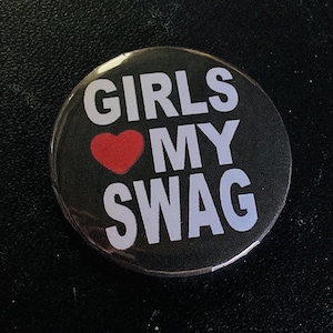 May include: A round, black button with the text "GIRLS MY SWAG" in white, with a red heart replacing the word "LOVE". The button is a fashion accessory.