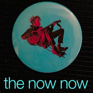 May include: A round, teal button featuring a red and black illustration of a person playing a guitar. The text "the now now" is printed in teal at the bottom of the button. The button has a glossy finish.