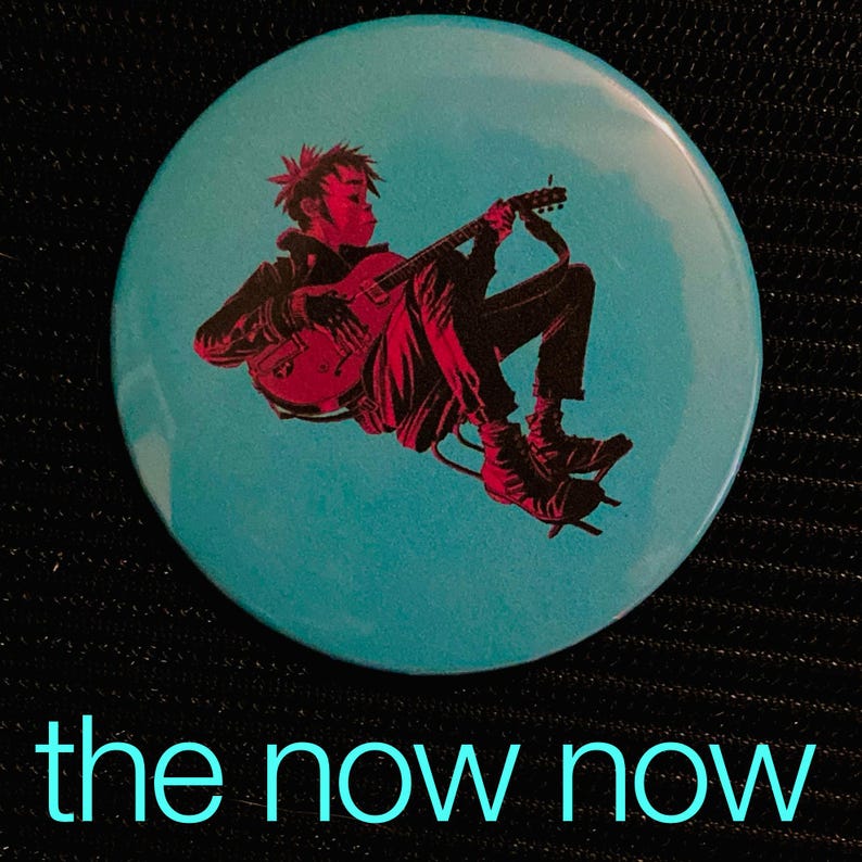 May include: A round, teal button featuring a red and black illustration of a person playing a guitar. The text "the now now" is printed in teal at the bottom of the button. The button has a glossy finish.