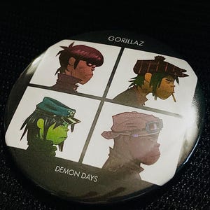 May include: A round button featuring four cartoon portraits of the band Gorillaz, with the text "GORILLAZ" at the top and "DEMON DAYS" at the bottom. The portraits are in a grid pattern, each with a different character.