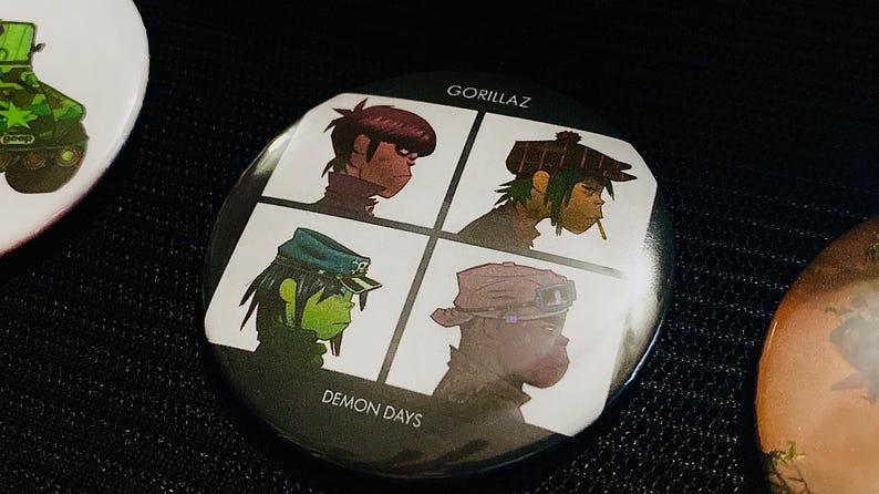 May include: A round button featuring four cartoon portraits of the band Gorillaz, with the text "GORILLAZ" at the top and "DEMON DAYS" at the bottom. The portraits are in a grid pattern, each with a different character.