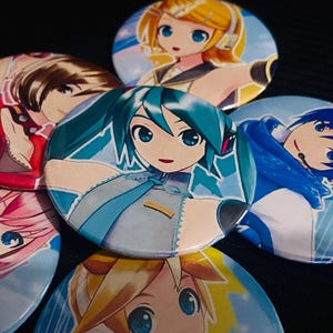 Op de afbeelding: Een verzameling ronde anime-personagebuttons. De buttons tonen kleurrijke illustraties van anime-personages met grote ogen en verschillende haarkleuren, waaronder blauw, geel, roze en bruin. De buttons zijn gerangschikt op een donkere ondergrond.