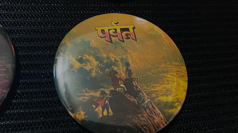 May include: Round button with a golden yellow background featuring an illustration of people on a mountain. The button has the word "पर्वत" in red letters at the top. The image has a vintage, adventure theme.