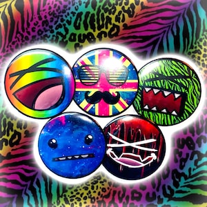 May include: A collection of five round, colorful buttons with unique designs. One button features a rainbow-colored open mouth, another has a Union Jack design with sunglasses and a mustache. Other buttons have monster and galaxy-themed designs.