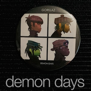 May include: A round button featuring the Gorillaz band's "Demon Days" album art. The design is divided into four squares, each with a different band member illustration. The button has the band's name and album title printed on it.