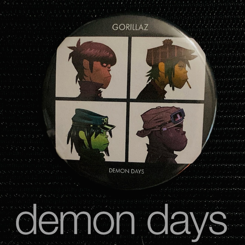 May include: A round button featuring the Gorillaz band's "Demon Days" album art. The design is divided into four squares, each with a different band member illustration. The button has the band's name and album title printed on it.