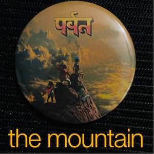 May include: Round button with a mountain scene, featuring figures on a rocky peak under a sunset sky. The button has the word "पर्वत" in gold lettering at the top and the words "the mountain" at the bottom.