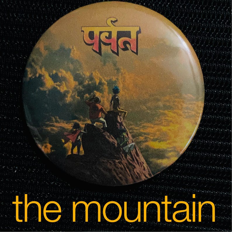 May include: Round button with a mountain scene, featuring figures on a rocky peak under a sunset sky. The button has the word "पर्वत" in gold lettering at the top and the words "the mountain" at the bottom.
