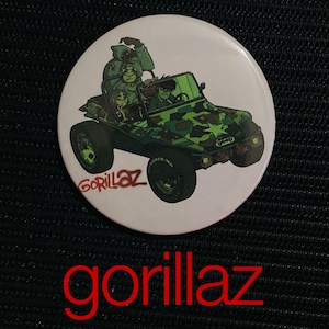 May include: A white button featuring a cartoon image of a green camouflage jeep with the band Gorillaz inside. The word "Gorillaz" is printed in red at the bottom of the image. The jeep has large black tires.