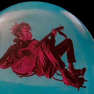 May include: A round, light blue button with a graphic of a person playing a guitar. The figure is in dark red, with a black jacket and pants. The button is set against a black background.