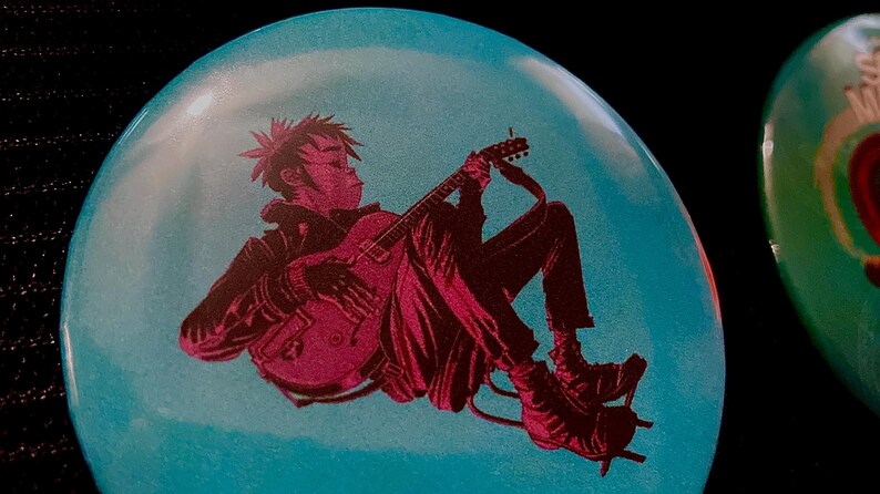 May include: A round, light blue button with a graphic of a person playing a guitar. The figure is in dark red, with a black jacket and pants. The button is set against a black background.