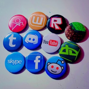 May include: A collection of round, colorful pinback buttons featuring logos of social media platforms and video games. The buttons include logos for YouTube, Facebook, Reddit, and Minecraft, among others. The buttons are arranged on a white surface.