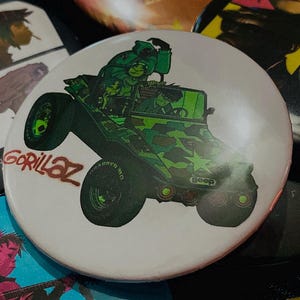 May include: A collection of Gorillaz band buttons. One button features a green camouflage jeep with the band's name. Other buttons display the band members in various styles. The buttons are round and colorful.