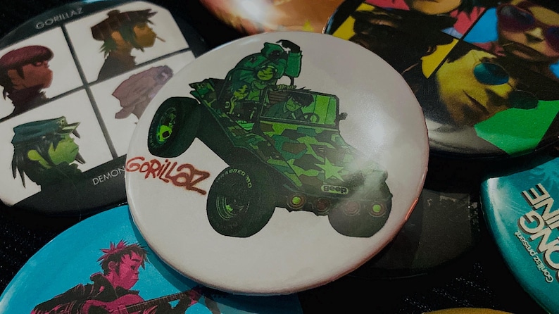 May include: A collection of Gorillaz band buttons. One button features a green camouflage jeep with the band's name. Other buttons display the band members in various styles. The buttons are round and colorful.