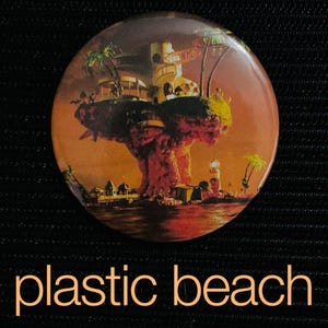 May include: Round button with a colorful illustration of a floating island with buildings, palm trees, and a lighthouse. The button has the words "plastic beach" in a light orange font at the bottom.