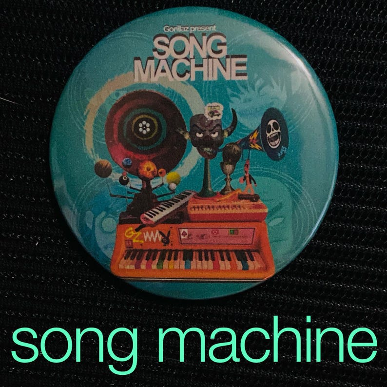 May include: A round, teal button featuring the text "Gorillaz present SONG MACHINE" and a colorful illustration of a keyboard, speakers, and a cartoon character. The words "song machine" are written in a green font at the bottom.