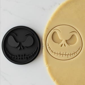 Può includere: Tagliapasta circolare nero con il volto di Jack Skellington, accanto a un pezzo di pasta con la stessa impronta. Il tagliapasta mostra gli occhi e il sorriso distintivi del personaggio. Ideale per la pasticceria.