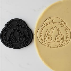 Può includere: Un tagliabiscotti nero a forma di volto di personaggio dei cartoni animati, accanto a un pezzo di pasta con lo stesso disegno impresso. Il tagliabiscotti ha un contorno dettagliato del volto, compresi occhi, bocca e capelli. La pasta mostra l'impronta del tagliabiscotti.