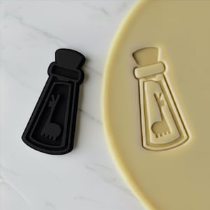 Può includere: Tagliapasta nero a forma di lama accanto a un biscotto con lo stesso disegno di lama impresso. Il tagliapasta è nero pieno e il biscotto è di colore marrone chiaro. Il disegno del lama è all'interno di un contorno a forma di bottiglia.