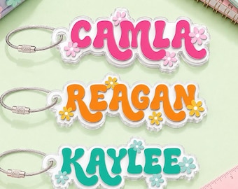 May include: Three acrylic keychains with metal wire loops, each featuring a name in a retro font. The top keychain is pink with white flowers and reads CAMLA, the middle is orange with yellow flowers reading REAGAN, and the bottom is teal with light blue flowers reading KAYLEE.