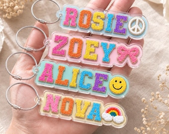 May include: Four rectangular acrylic keychains with metal wire loops. Each features colorful, textured varsity-style letters spelling the names ROSIE, ZOEY, ALICE, and NOVA, accompanied by a peace sign, a bow, a smiley face, and a rainbow icon.