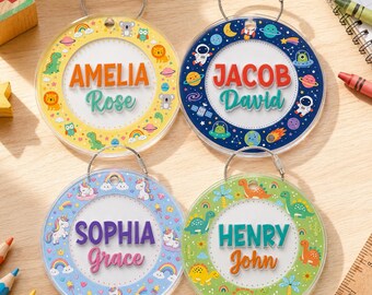May include: Four circular acrylic bag tags with metal wire loops. Each tag features a white center with a personalized name in colorful fonts, surrounded by a patterned border: yellow with animals, dark blue with space, light blue with unicorns, and green with dinosaurs.