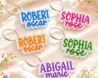 May include: Five custom acrylic keychains with silver wire loops. Each features a name in block letters above a second name in cursive script. Colors include blue, pink, orange, green, and purple, all set against a clear, scalloped-edge base.