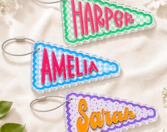 May include: Three triangular acrylic keychains with scalloped edges and metal wire loops. The top keychain is green checkered with the pink name HARPER. The middle is blue checkered with the pink name AMELIA. The bottom is purple dotted with the orange name Sarah.