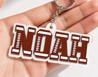 May include: A custom acrylic keychain featuring the name NOAH in brown block letters with white accents. Each letter is designed to resemble a football, complete with white laces and stripes. The keychain includes a silver metal ring and chain.